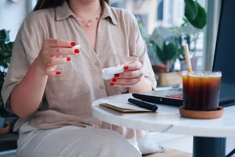 A woman handling wireless earbuds beside iced coffee and a laptop at a cafe table.
