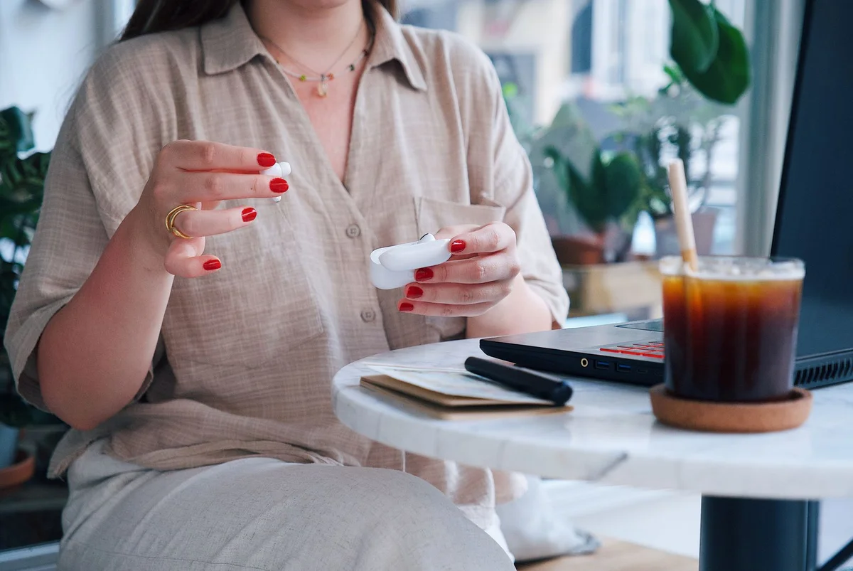 A woman handling wireless earbuds beside iced coffee and a laptop at a cafe table.